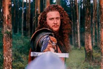 Ein junger Mann mit langen Locken und Lederkleidung steht in einem Kiefernwald.